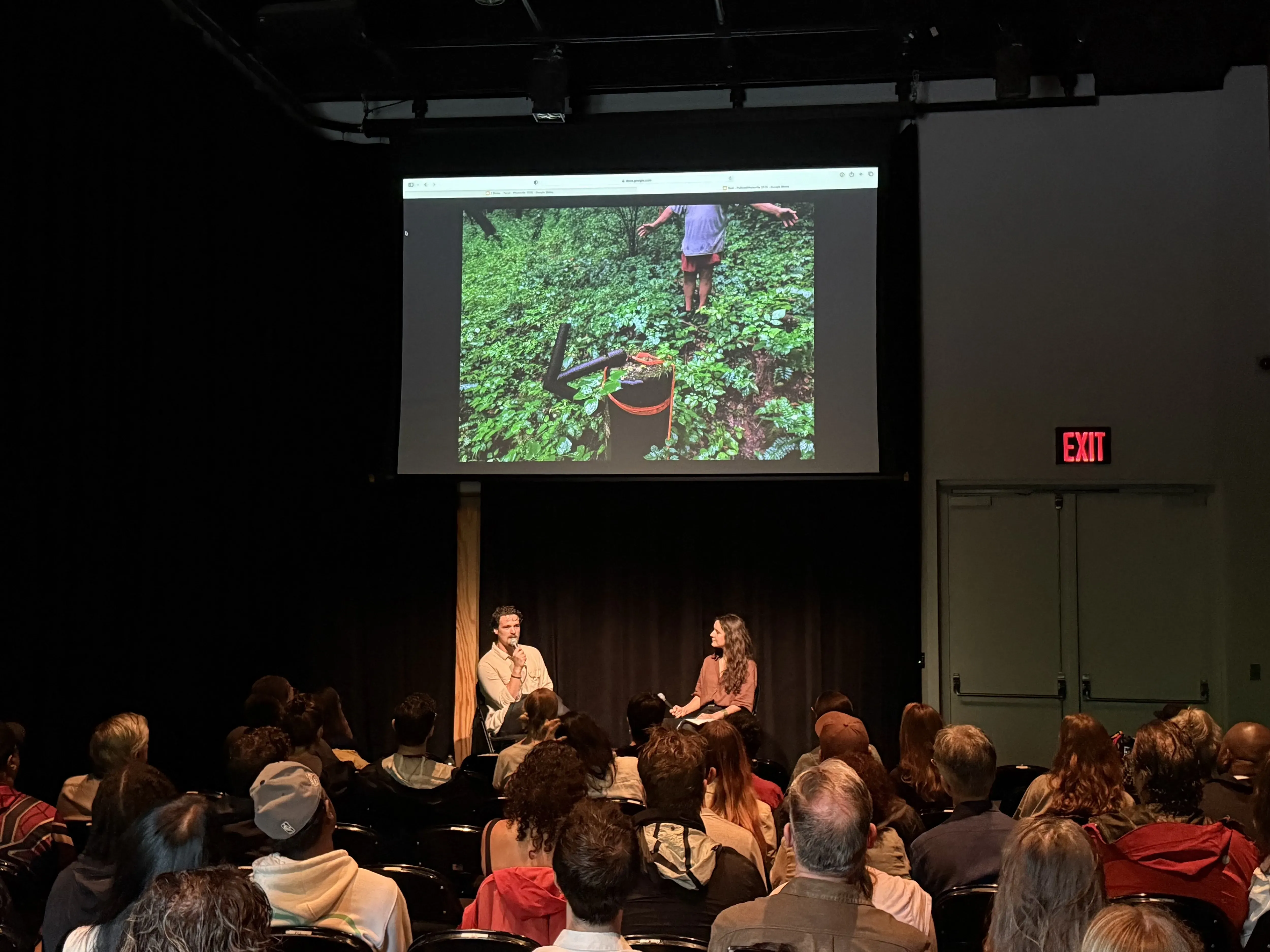 Two people sit on a stage speaking into microphones under a screen displaying reporting photos.